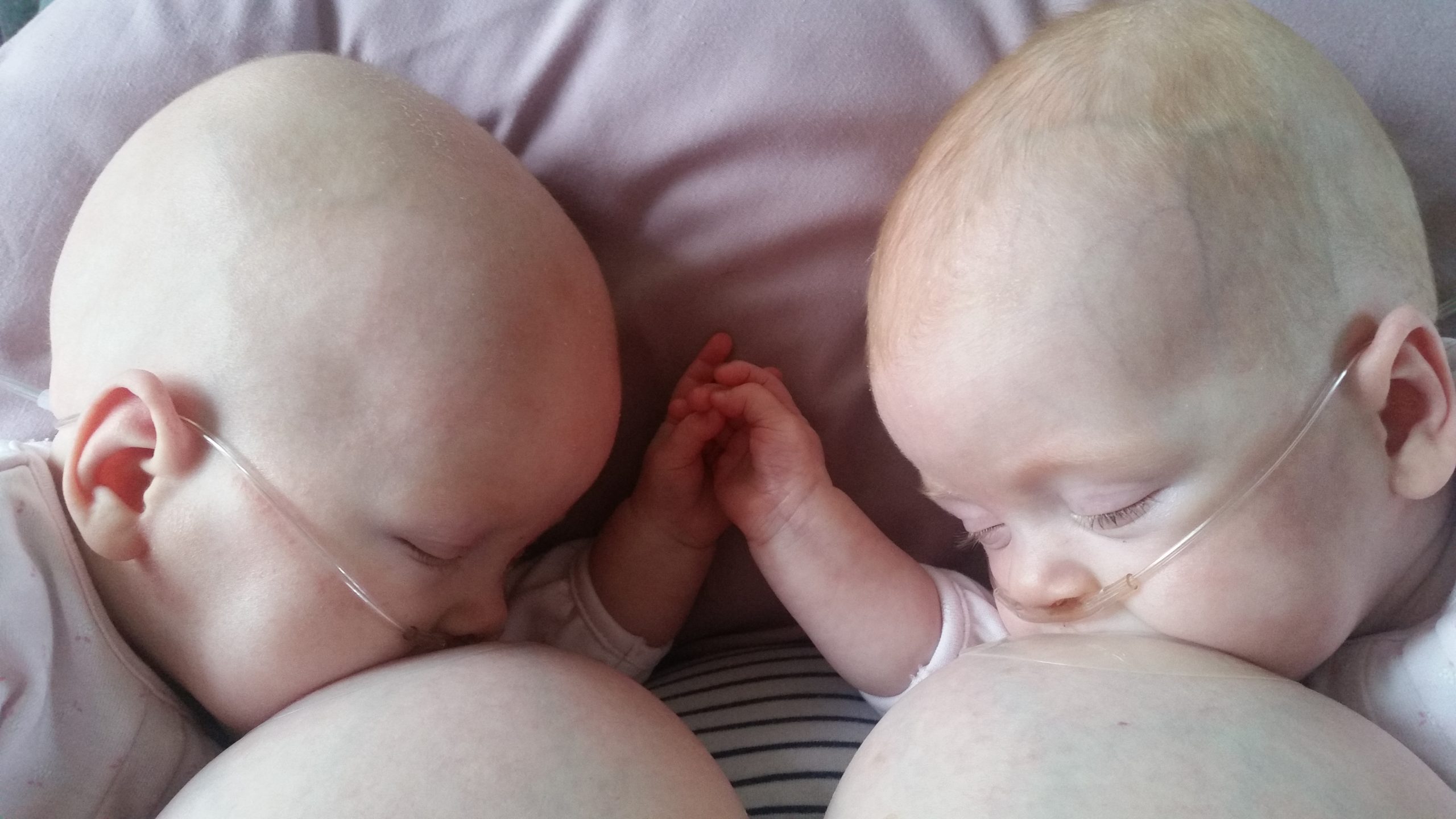 Twin babies hold hands while feeding from a breast each.