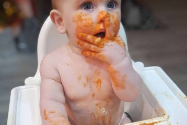 A baby sits in a white highchair, eating tomato pasta. The pasta sauce is all over their face, body and high chair.